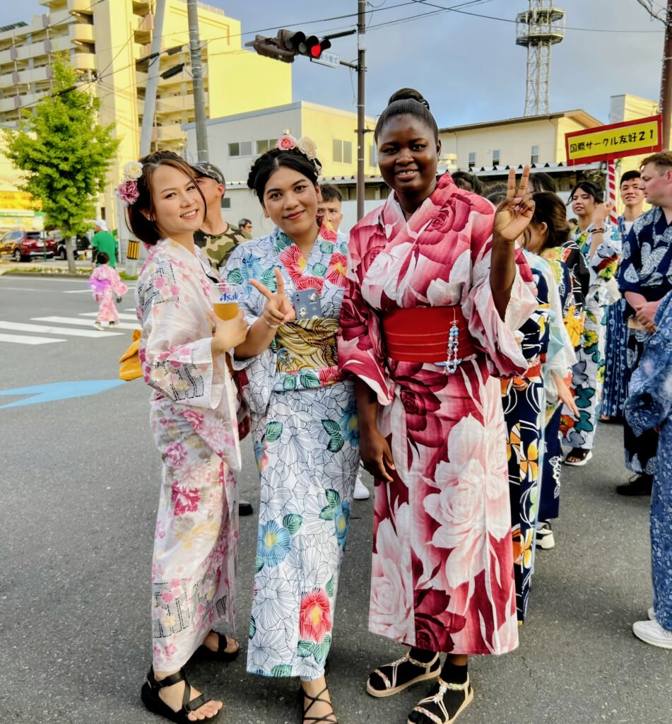 Smiling group of people wearing colorful yukata at a Japanese summer festival, posing with peace signs.
