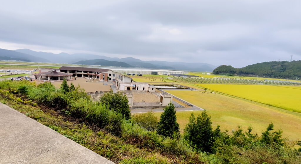 Okawa School Tsunami Memorial – Stories of Hope and Resilience Wide view of Okawa Elementary School ruins in Ishinomaki, surrounded by fields and mountains, preserved as a tsunami memorial site.